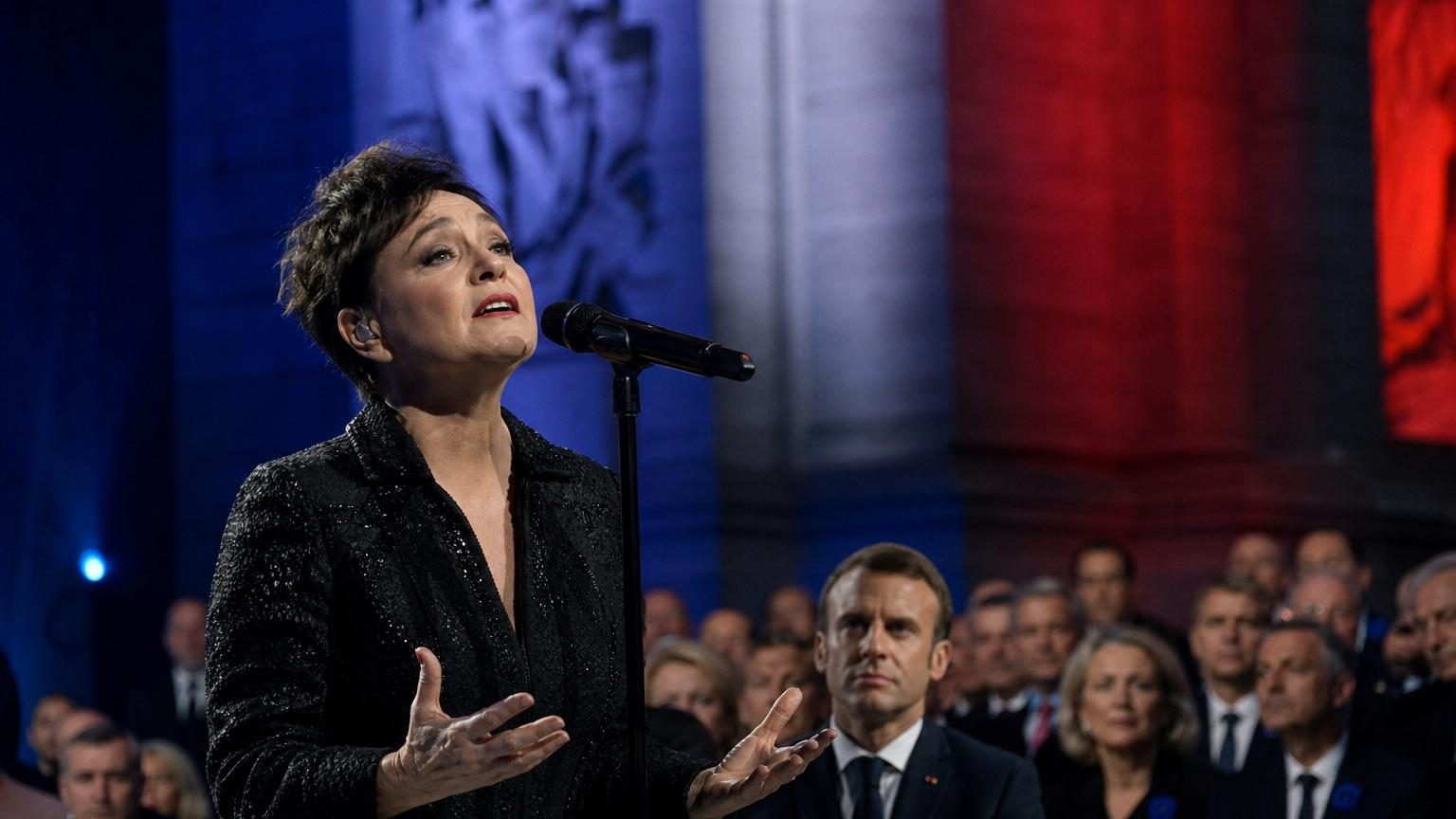 Catherine Ringer chante sur scène lors d'une cérémonie officielle en présence d'Emmanuel Macron, visages graves, lumière solennelle.