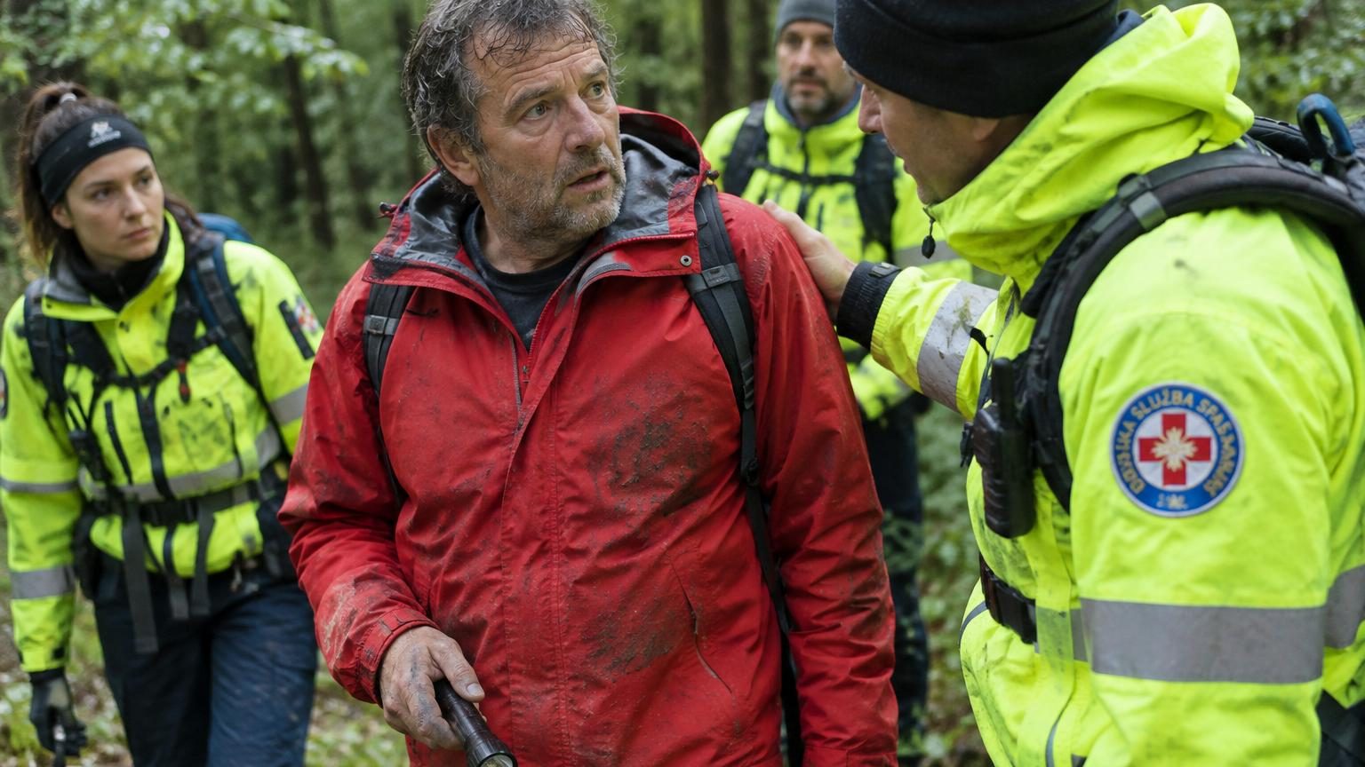 Un homme en vêtements de sauvetage marche parmi les secouristes dans une forêt sombre, cherchant la personne qui est en réalité lui-même.